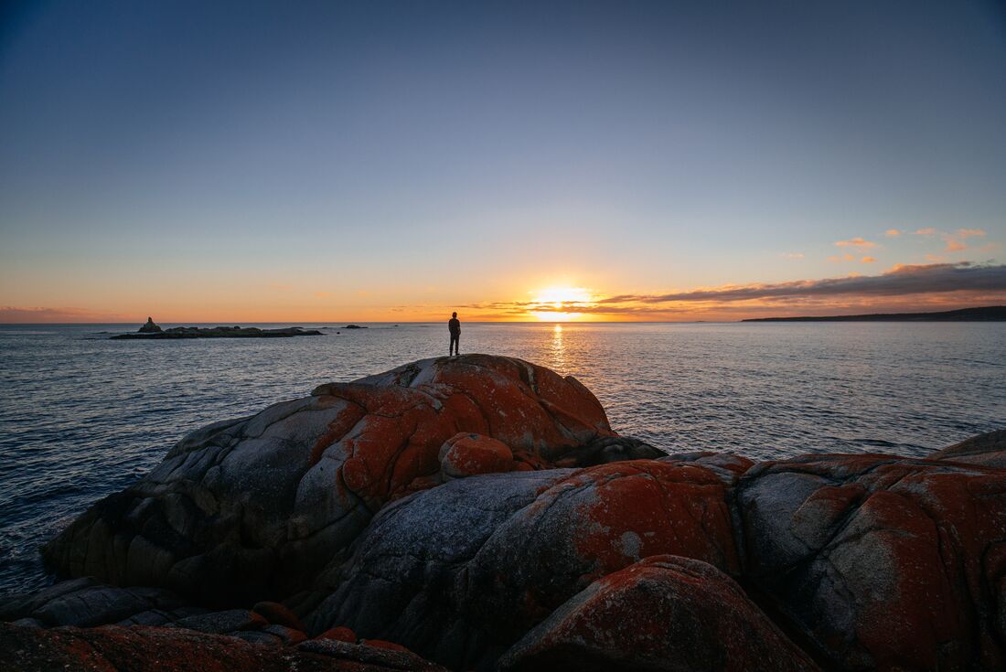 Intrepid traveller stands on Bay of Fires boulder shore looking out at the sunset over the Tasman Sea