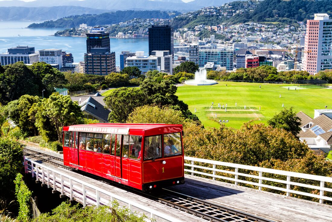 Red tram on tracks in Wellington, with cityscape and a cricket field in the background, New Zealand
