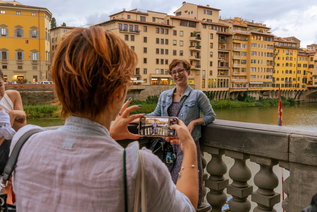Leader takes a photo of traveller standing on a bridge in Florence with yellow buildings in the background, Italy