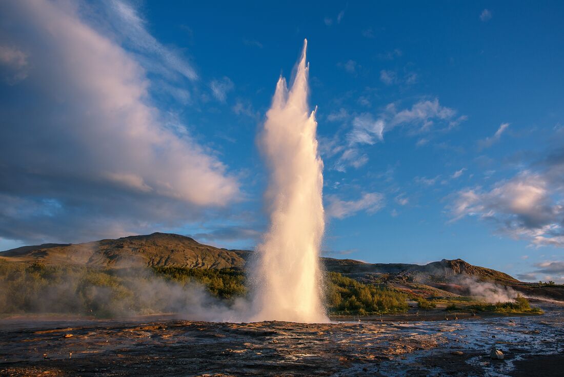 Strokkur Geyser erupting during sunset with blue sky and white fluffy clouds